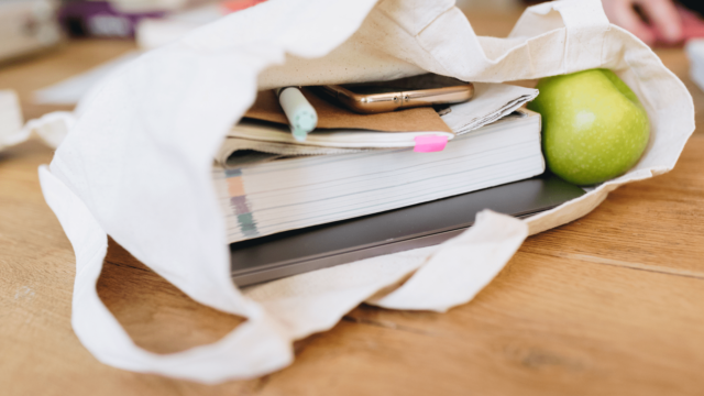 Un sac en tissu dans lequel il y a une pomme, un livre, un ordinateur, un carnet et un téléphone.