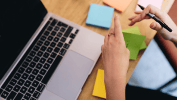 Une femme écrit quelque chose sur un post it devant son ordinateur.