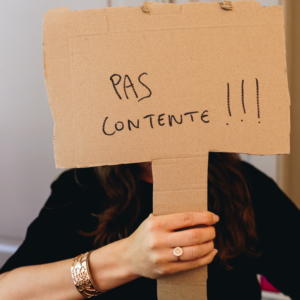 Une personne cache son visage avec une pancarte en carton sur laquelle il est marqué "Pas contente".
