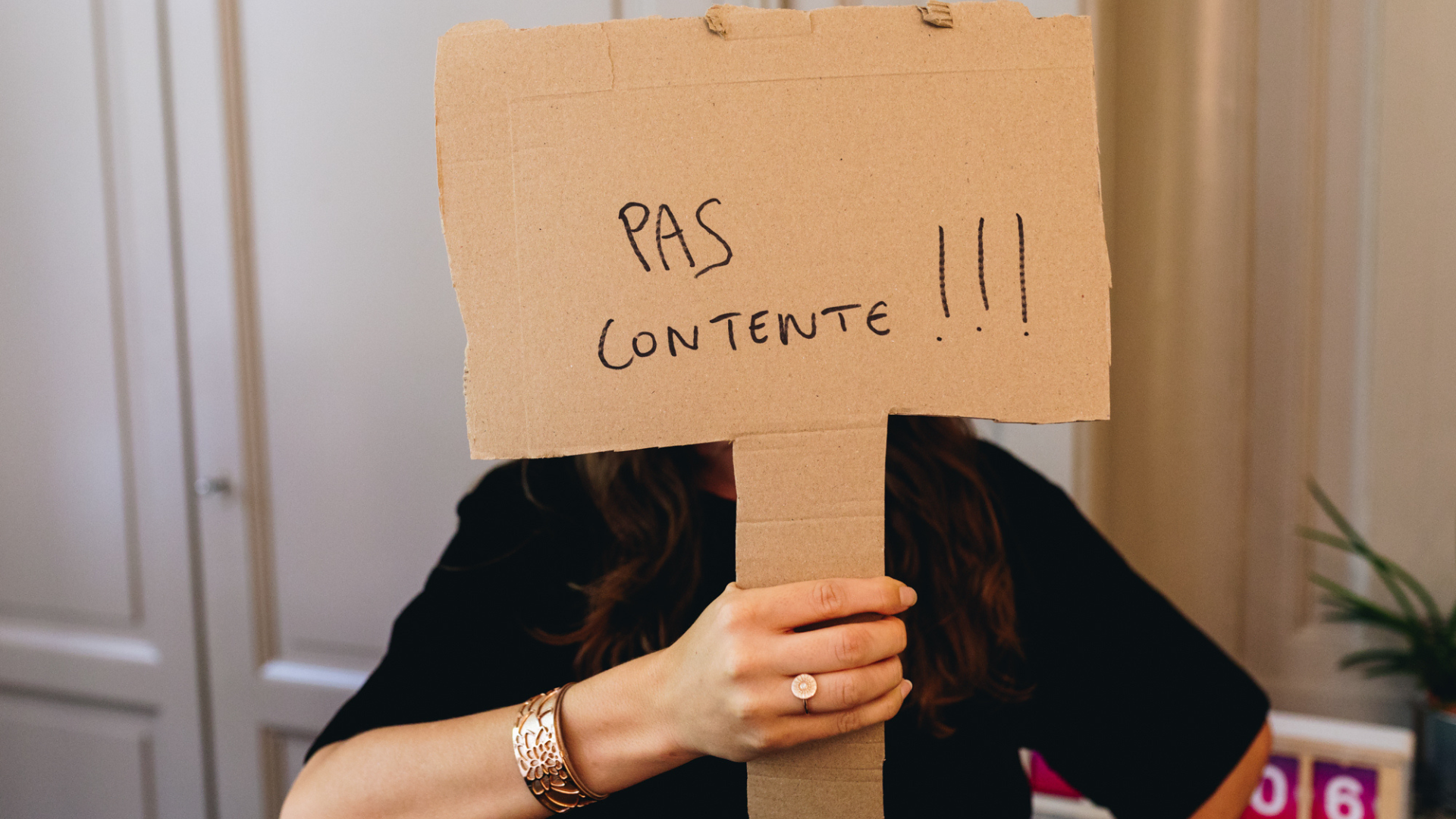 Une personne cache son visage avec une pancarte en carton sur laquelle il est marqué "Pas contente".