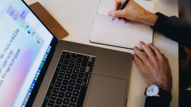 une personne qui écrit sur un carnet, un ordinateur sur la table