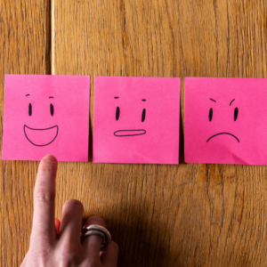Sur une table en bois, trois post it représentent trois émotions : la joie, neutre, la colère. Un doigt pointe le post it joyeux.
