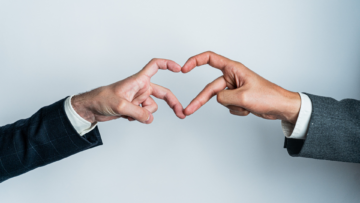 Deux hommes en costume forment un cœur avec leurs doigts, sur un fond gris.