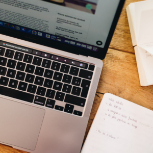 Un ordinateur ouvert est posé sur une table, à côté, il y a un carnet de notes ouvert et un stylo.