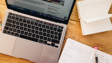 Un ordinateur ouvert est posé sur une table, à côté, il y a un carnet de notes ouvert et un stylo.