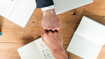 Deux hommes se cognent les poings au dessus d'une table encombrée de carnets de notes.
