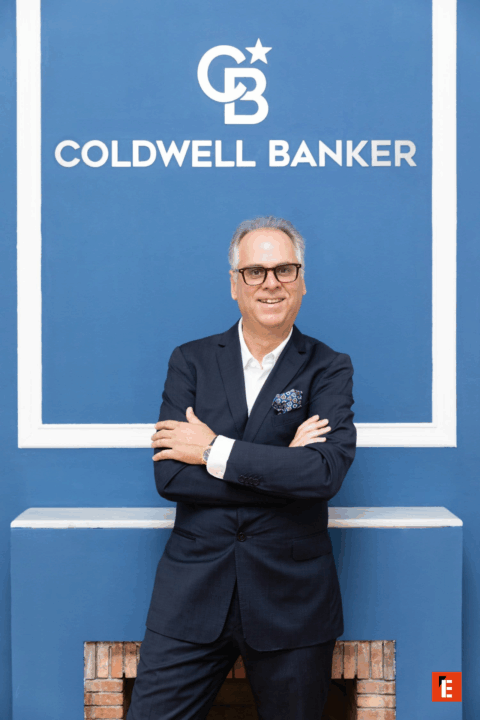 Hombre sonriente frente al logo de Coldwell Banker.
