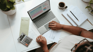 Close up of a person's hands on a computer. A calculator is next to the computer along with sheets of paper and a coffee.