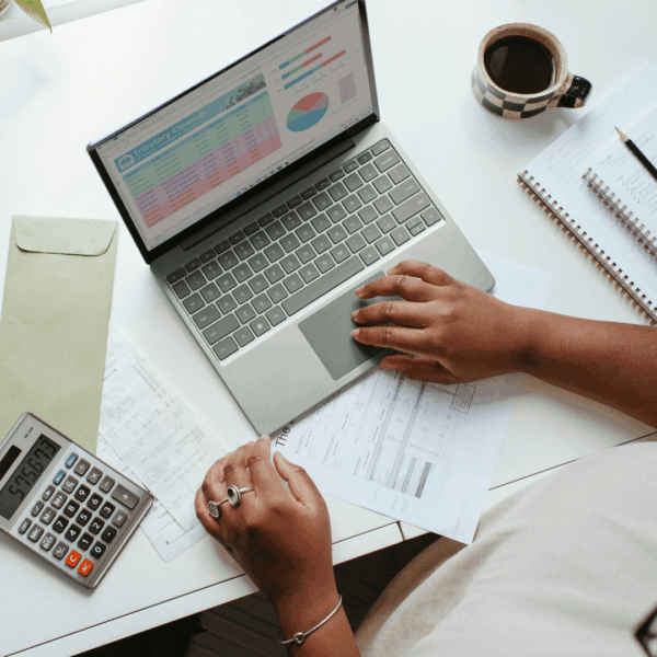 Close up of a person's hands on a computer. A calculator is next to the computer along with sheets of paper and a coffee.