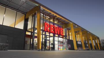 beleuchtete rewe glasfassade am abend mit holzstützen