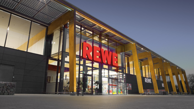 beleuchtete rewe glasfassade am abend mit holzstützen
