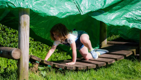 child crawling on wooden play bridge under green tarp